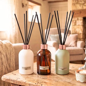 May include: Three reed diffusers in white, brown, and green glass bottles with copper-coloured collars. Each bottle has black reeds and a label that reads "SCENT GROVE". The diffusers are on a wooden table.