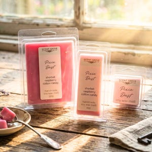 May include: Three pink soy wax melts in clear plastic packaging. The largest one reads "Pixie Dust" and "sherbet raspberry cotton candy." The smallest one reads "soy wax melt." The packaging is on a wooden surface.