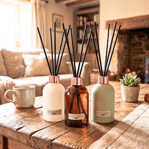 May include: Three reed diffusers in cream, amber, and sage green bottles with copper-coloured collars and black reeds. Each bottle has a "SCENT GROVE" label. The diffusers are on a wooden table with a mug and a small potted succulent.