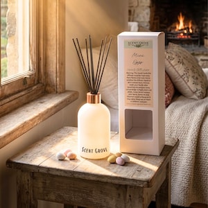 May include: A white glass reed diffuser with a copper-coloured collar and black reeds, labelled "Scent Grove." The box reads "Mini Eggs reed diffuser." Pastel-coloured candies sit on a rustic wooden table near a window.
