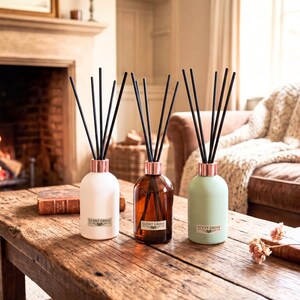 May include: Three reed diffusers in white, amber, and sage green, with black reeds and copper-coloured collars. The bottles are labelled "SCENT GROVE" and are displayed on a rustic wooden table.