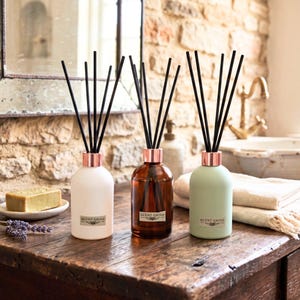 May include: Three glass reed diffusers in white, brown, and green, each with a rose gold collar and black reeds. The bottles, labelled "SCENT GROVE", sit on a wooden surface. A bar of soap and lavender are also visible.