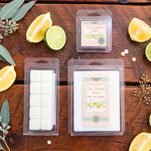 May include: Two clear plastic clamshell containers holding white wax melts. One container holds ten small square cubes, the other a large rectangular block. Labels read Scent Grove Sun Washed Linen with lemon, lime, and eucalyptus graphics.