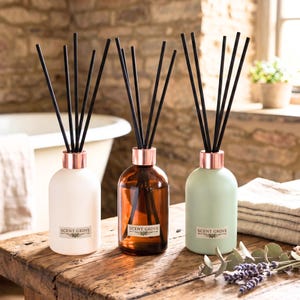 May include: Three reed diffusers on a wooden surface. The diffusers are in white, amber, and green glass bottles with copper-coloured collars and black reeds. Each bottle has a label that reads "SCENT GROVE".