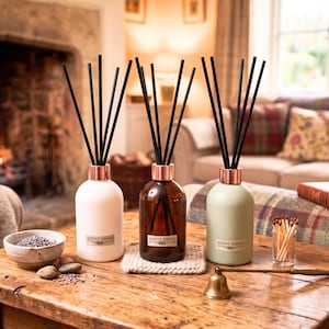 May include: Three reed diffusers in white, brown, and sage green, with black reeds and copper-coloured collars. The bottles are labelled "SCENT GROVE." A small bowl of lavender, matches, and a bell are on a wooden table.
