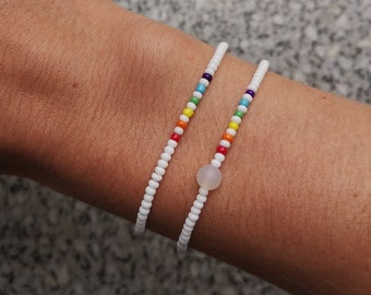 May include: Two white beaded bracelets with a rainbow of colored beads on one bracelet and a single white bead on the other.
