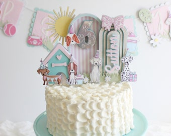 May include: A white cake with buttercream frosting decorated with a dog-themed cake topper. The topper includes a dog house, a sun, a number six, a bow, the name "Sutton", and several dogs. The cake is on a light blue cake stand.