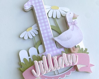 May include: A decorative cake topper featuring a large number 1 with a pink gingham pattern. A pink banner displays the name "Jillian" in cursive. A white goose with a pink bow and daisies complete the design.