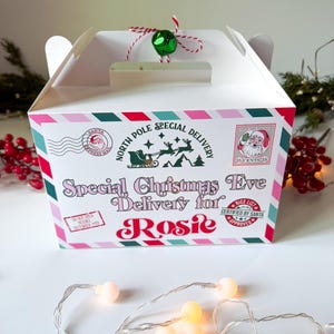 May include: A white Christmas-themed gift box with a handle, decorated with pink, red, and green stripes. The box features text that reads "Special Christmas Eve Delivery for Rosie" and a Santa Claus stamp. A green bell hangs from the top.