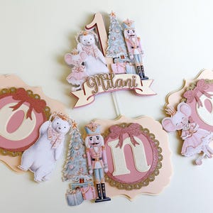 May include: A collection of party decorations featuring a Nutcracker theme. Includes a cake topper with the number 1, a banner with the name "Milani", and decorative cutouts of a polar bear, mouse, and Nutcracker soldier. The color scheme is pink, gold, and white.