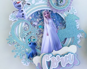 May include: A blue and white cake topper featuring a Frozen character, Elsa, with a sparkly blue horse, a blue lizard, and a snowman. The topper also includes a glittery circle with the number 5 and the name "Cora" in a blue and white font.
