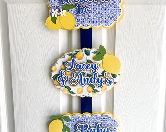 May include: Three blue and white patterned signs with yellow lemons and white flowers. The top sign says "Welcome to", the middle sign says "Lacey & Andy's", and the bottom sign says "Baby Shower".