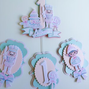 May include: A festive cake topper and decorative cutouts featuring a pastel color scheme. The cake topper includes a number 1, a gingerbread man, a Christmas tree, and a nutcracker. The cutouts include a nutcracker, a number 1, and an elf. The name "Allison" is also present.