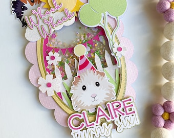 May include: A pink and gold cake topper with a white guinea pig wearing a party hat. The topper says "Wheek Wheek" and "Claire turns six".