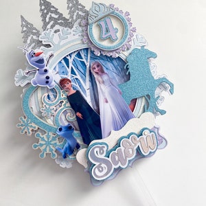 May include: A blue and white cake topper featuring characters from the movie Frozen. The topper includes Elsa, Anna, Olaf, and a blue unicorn. The topper also includes a number 4 and the name "Savri".