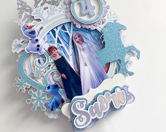 Könnte beinhalten: Ein blauer und weißer Tortenaufsatz mit Figuren aus dem Film Frozen. Der Aufsatz enthält Elsa, Anna, Olaf und ein blaues Einhorn. Der Aufsatz enthält auch die Zahl 4 und den Namen "Savri".