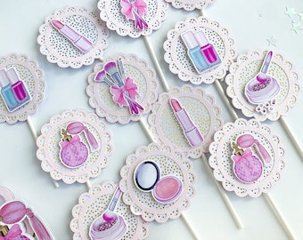 May include: A set of 10 pink and gold cupcake toppers featuring various makeup and beauty items, including lipstick, nail polish, perfume bottles, brushes, and eyeshadow palettes. The toppers are decorated with white lace and gold glitter.
