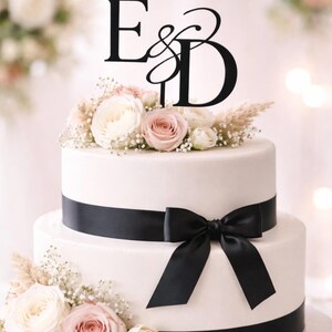 May include: A two-tiered white wedding cake, decorated with a black ribbon and bow. The cake is adorned with fresh flowers in white and pink. A black cake topper displays the letters "E & D".