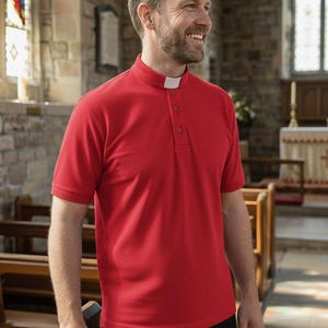 Op de afbeelding: Een rood poloshirt met korte mouwen en een opstaande kraag met een witte priesterboord-inzet bij de hals. Het shirt heeft een gedeeltelijke knoopsluiting met drie bijpassende rode knopen en een textuur in de stof, gedragen met een zwarte broek.