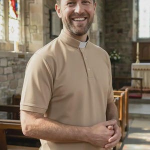 Op de afbeelding: Een beige poloshirt met korte mouwen, voorzien van een opstaande kraag en een wit priesterboordje bij de halslijn. Het shirt heeft een knoopsluiting, een rechte zoom met zijsplitten en een gladde stoftextuur voor formele religieuze kleding.
