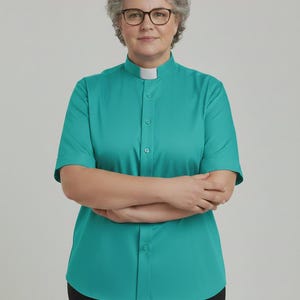 May include: A short-sleeved teal clergy shirt featuring a white clerical collar insert, a button-down front placket, and a straight hemline. The shirt is worn with black trousers and is paired with dark-rimmed glasses and short gray hair.