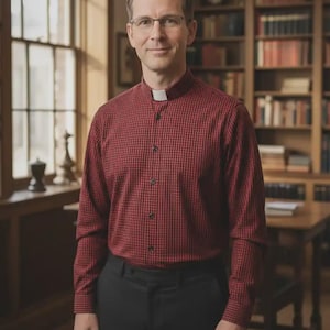 May include: A man wearing a red and black checkered shirt with a white collar and black pants. The man is wearing glasses and standing in front of a bookshelf.