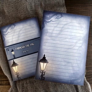 May include: Two memo pads with a spooky theme. The pads have a blue and gray gradient background with a street lamp illustration. The text "THROUGH THE FOG" is printed on the cover. The pads are lined and rest on a gray fabric.