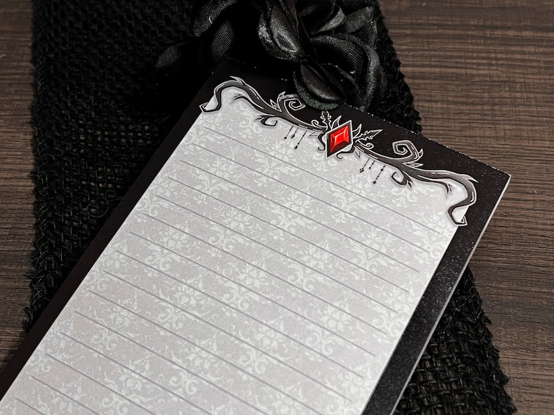 May include: A black and white notepad with a decorative border and a red gem. The notepad has lined pages with a floral pattern. The notepad is resting on a black mesh fabric.