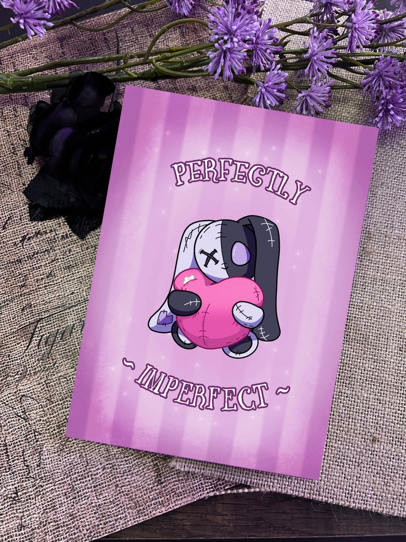 May include: A pink and purple card with a cartoon illustration of a stitched-together plush toy holding a pink heart. The text "PERFECTLY IMPERFECT" is written in black on the card.