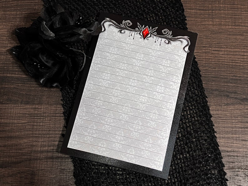 May include: A lined notepad with a black border and a decorative top design featuring a red gem. The notepad has a white background with a subtle floral pattern. A black fabric flower is in the upper left corner.