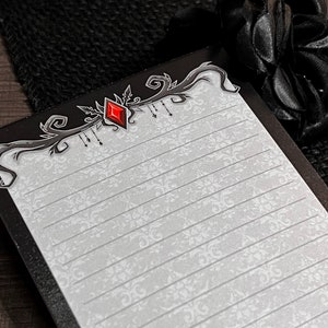 May include: A lined notepad with a decorative black border featuring a red diamond-shaped gem. The notepad has a white background with a subtle floral pattern. A black fabric and a black rose are in the background.