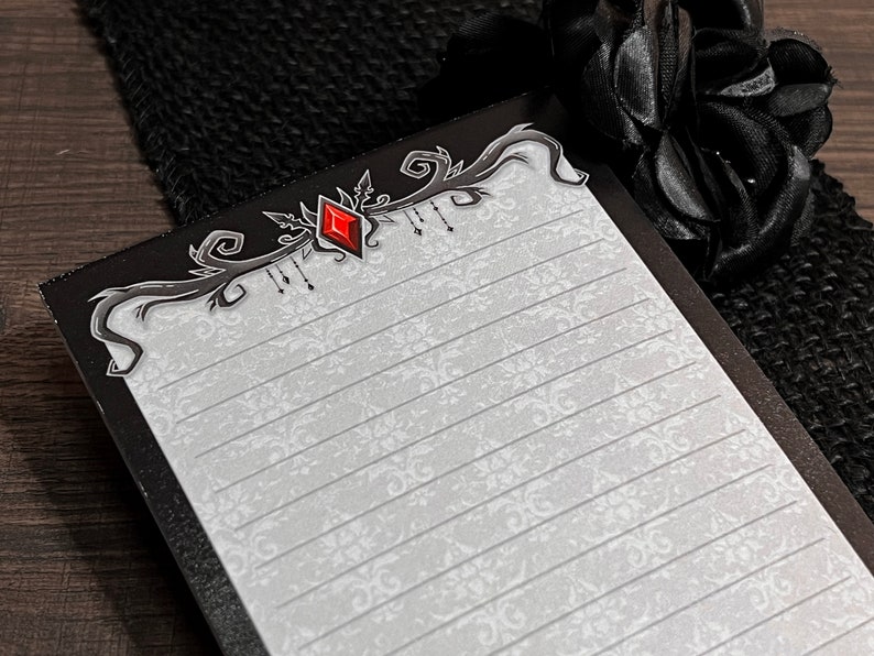 May include: A lined notepad with a decorative black border featuring a red diamond-shaped gem. The notepad has a white background with a subtle floral pattern. A black fabric and a black rose are in the background.