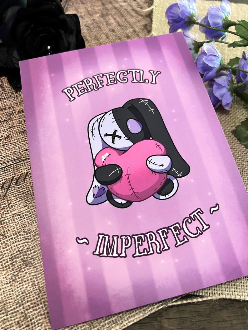 May include: A pink and purple card with a cartoon illustration of a stitched plush toy holding a pink heart. The text on the card reads "Perfectly Imperfect."