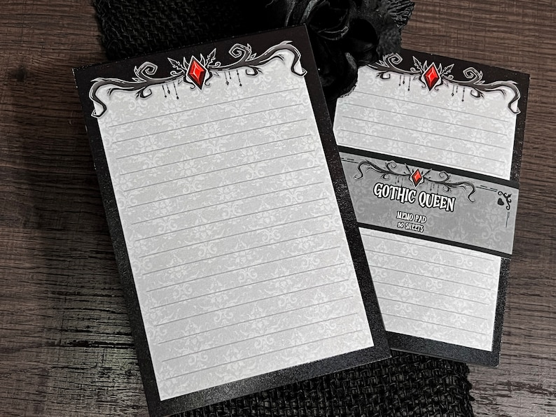 May include: Two memo pads with a gothic design. The pads have a black border and a white background with a subtle pattern. The top of each pad features an ornate design with a red gem. One pad has the text "GOTHIC QUEEN MEMO PAD 50 SHEETS."