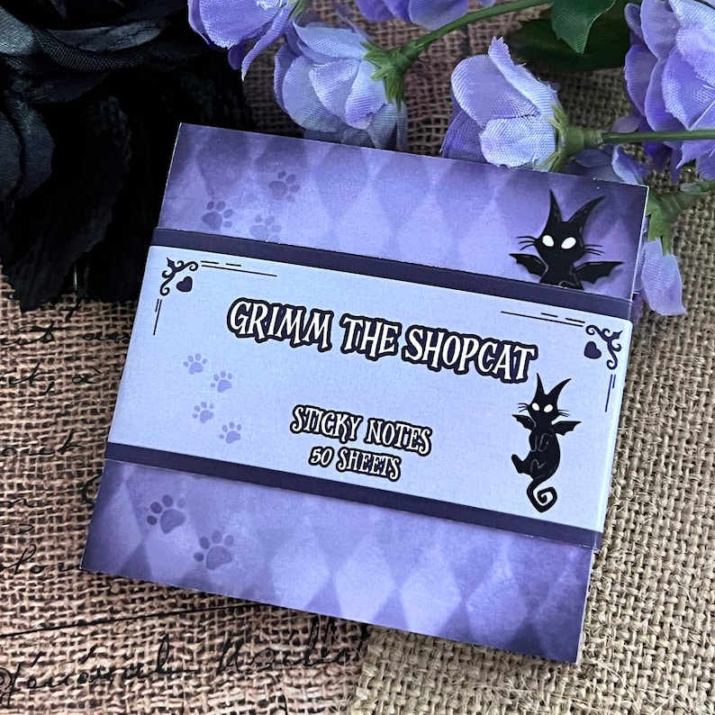 May include: A stack of "Grimm the Shopcat" sticky notes with a purple diamond pattern background. The label features the text "Grimm the Shopcat Sticky Notes 50 Sheets" with a black cat illustration and paw prints.