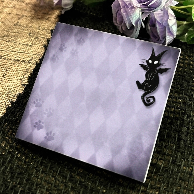 May include: Square notepad with a light purple diamond pattern and paw prints. A black cat with bat wings is in the corner. The notepad is on a textured surface with purple flowers in the background.