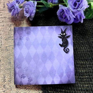 May include: Square purple notepad with a diamond pattern and paw prints. A black cat with bat wings is on the upper right corner. The notepad is surrounded by purple flowers.