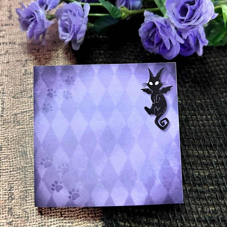 May include: Square purple notepad with a diamond pattern and paw prints. A black cat with bat wings is on the upper right corner. The notepad is surrounded by purple flowers.