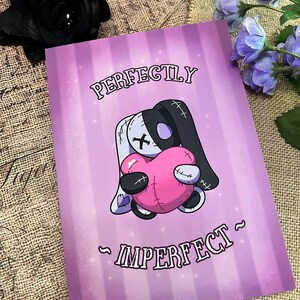 May include: A purple and white card with a cartoon illustration of a stitched plush toy holding a pink heart. The text "Perfectly Imperfect" is written on the card.