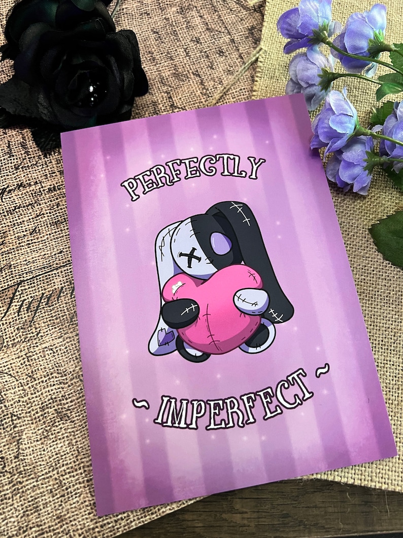 May include: A purple and white card with a cartoon illustration of a stitched plush toy holding a pink heart. The text "Perfectly Imperfect" is written on the card.