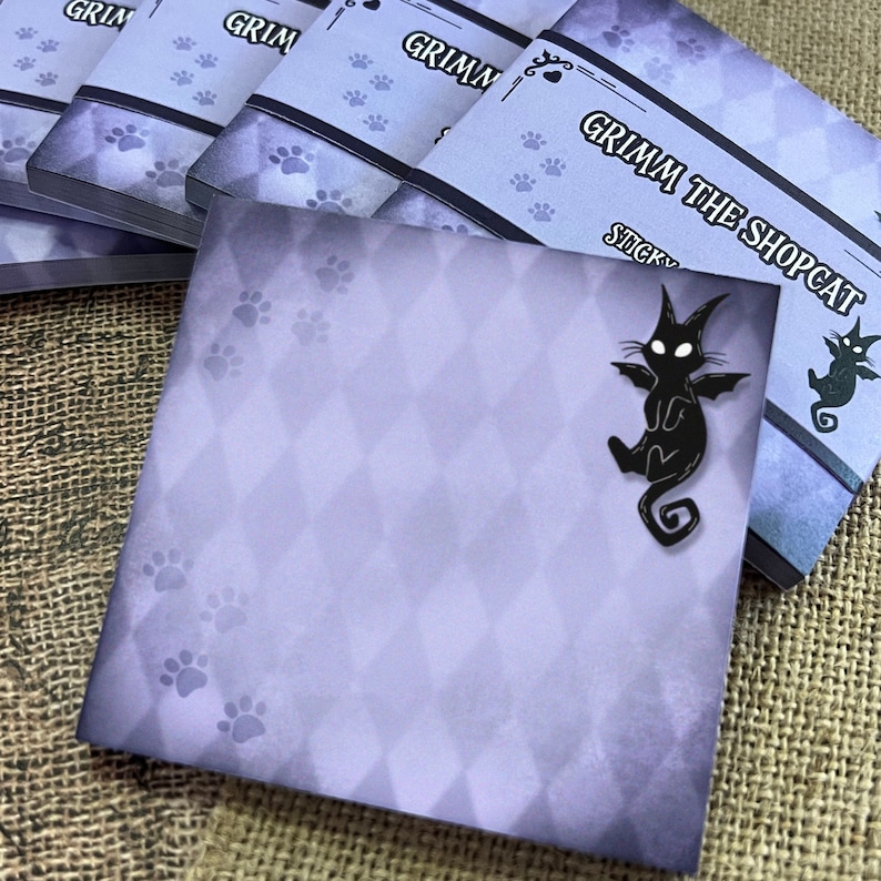 May include: Stack of sticky notes with a light purple diamond pattern and paw prints. The notes feature a black cat graphic with the text "Grimm the Shopcat". The background is a burlap texture.