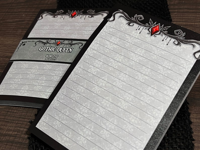 May include: Two memo pads with a black border and lined pages. The pads have a decorative floral pattern and the words "GOTHIC QUEEN" on the cover. The top border features a red gem design.