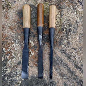 May include: Three wood chisels with black blades and wooden handles. The handles are light brown, and the blades are dark grey. The chisels are arranged vertically on a wood surface covered in wood shavings.
