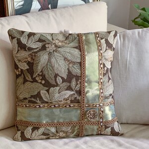 May include: A decorative throw pillow with a brown and green floral pattern. The pillow has a green satin ribbon and gold trim.