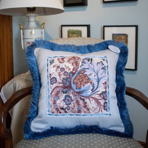 May include: A blue and white throw pillow with a floral design and blue fringe. The pillow has a white background with a floral pattern in shades of pink, blue, and green. The fringe is a deep blue color.