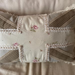May include: A rectangular throw pillow featuring a Union Jack pattern constructed from beige and brown striped fabric, light brown gingham fabric, and a central cream panel with a small pink rose floral print, all accented with white lace trim and fringe.