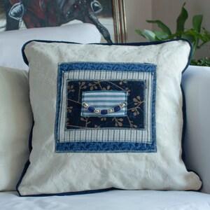 May include: A white pillow with a blue and white patterned fabric insert. The insert features a blue and white striped fabric with a row of blue beads.
