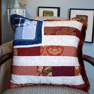 May include: A red, white and blue patchwork cushion with a fringed edge. The cushion is shaped like an American flag and is made of different fabrics.