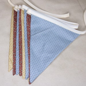 May include: A fabric bunting with alternating triangular flags in various patterns. The flags include a light blue dotted pattern, a brown floral pattern, and a cream floral pattern. The bunting is strung on a white ribbon.