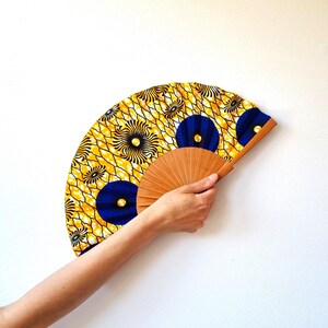 May include: A hand fan featuring a vibrant yellow and white geometric pattern with circular blue accents. The fan is constructed with a light brown wooden frame that radiates from the center, supporting the patterned fabric canopy in a semi-circle.